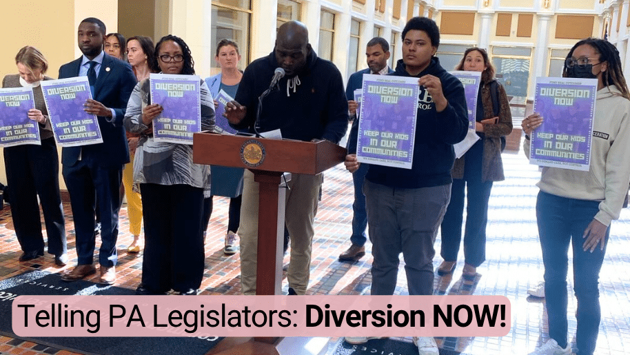 Photo: people in Harrisburg's Capitol Building standing around Jarue at lectern, holding signs that say Diversion Now. Text: Telling PA Legislators: Diversion NOW!